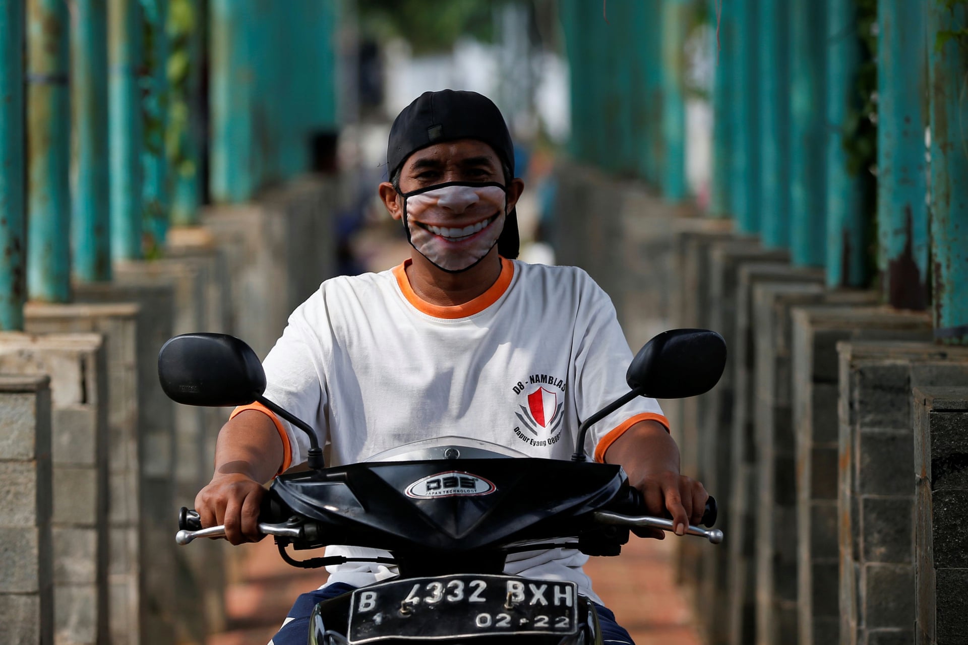 Indoneziyaning Jakarta shahrida kulib turgan niqob taqqan erkak mototsiklda harakatlanmoqda.