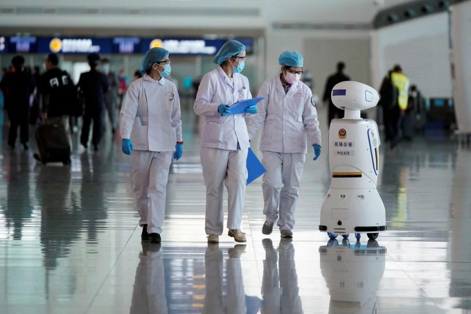 Uxan Tyanxe xalqaro aeroportida robot politsiyachi yonidan o‘tib ketayotgan tibbiyot xodimlari.