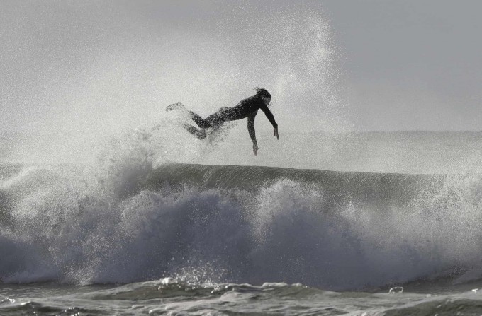 Янги Зеландиянинг Крайстчерч шаҳридаги Нью-Брайтон-Бич пляжида доскада тўлқинларни тутиб юрган сёрфер.