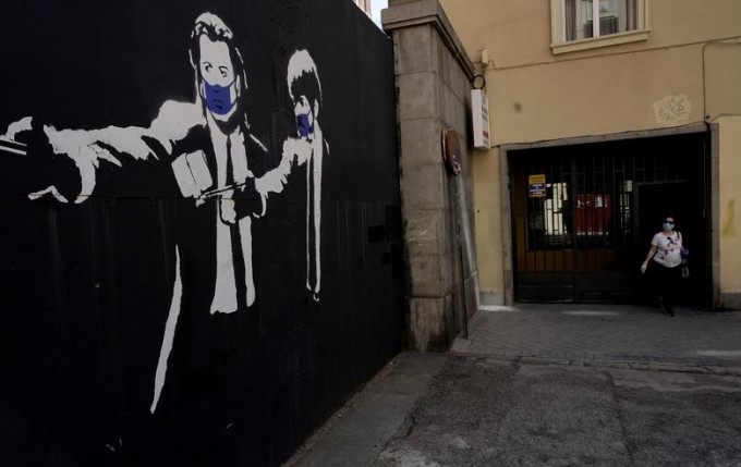 Испаниянинг Мадрид шаҳрида актёрлар Жон Траволта ва Семюел Л. Жексон ҳимоя ниқобини кийиб олган ҳолда тасвирланган граффити ёнида юрган аёл.