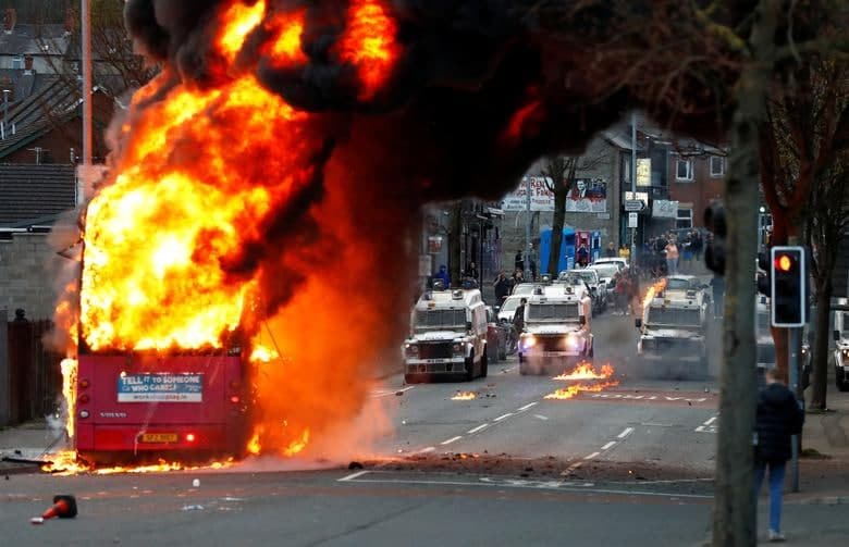 Shimoliy Irlandiyaning Belfast shahrida norozilik namoyishlari davom etayotgan paytda o‘g‘irlab, o‘t qo‘yib yuborilgan avtobus qarshisida turgan politsiya avtomobillari.