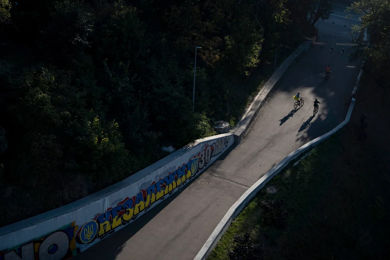 Украина байроғини ёпинган ўсмирлар Мустақиллик куни учун чизилган «Независими 30» ёзувли граффити ёнидан велосипедда ўтмоқда.