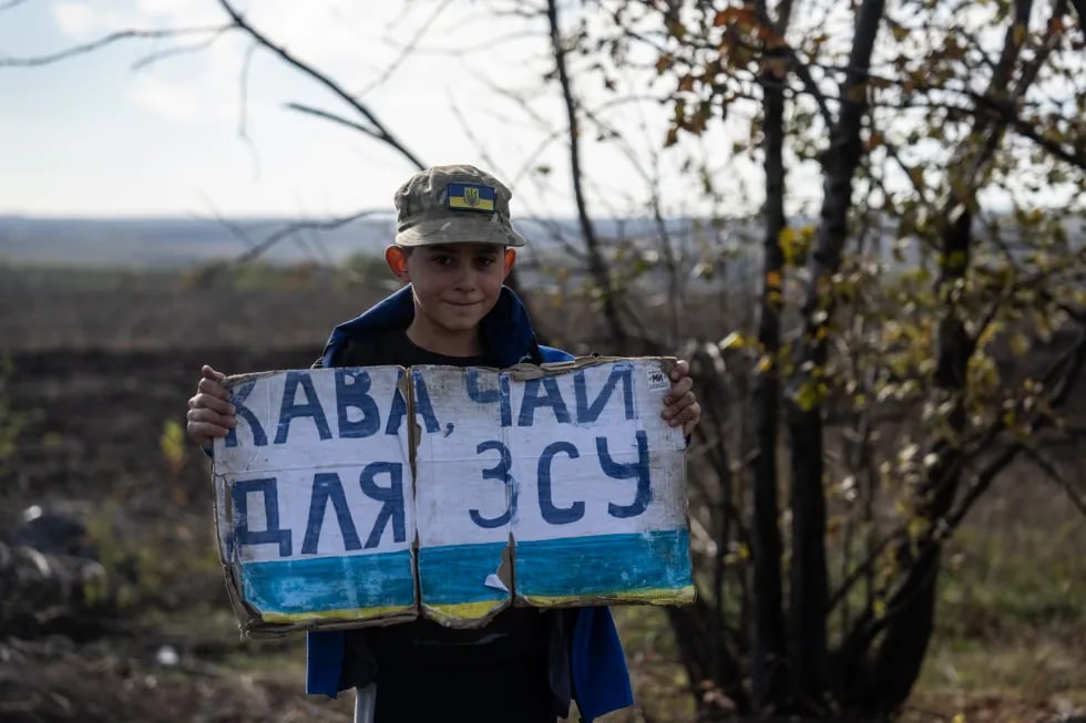 Dubovoye qishlog‘i yaqinidagi yo‘l bo‘yida “Ukraina Qurolli kuchlari uchun qahva, choy” yozuvli plakat ko‘targan bola.